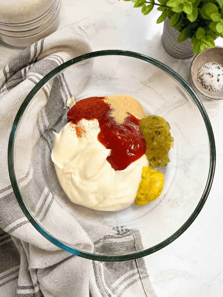 Ingredients for homemade burger sauce in a glass mixing bowl including mayonnaise, ketchup, yellow mustard, pickle relish, paprika, and garlic powder on a white countertop 