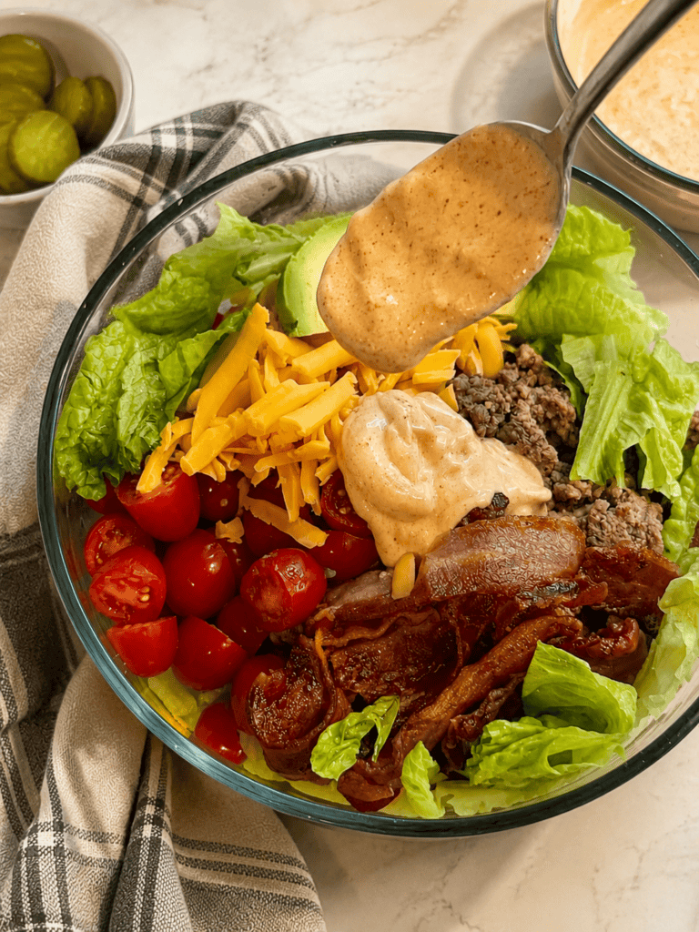 Loaded burger bowl topped with seasoned ground beef, bacon, cheddar cheese, avocado, lettuce, tomatoes, and burger sauce being drizzled from a spoon above the bowl.