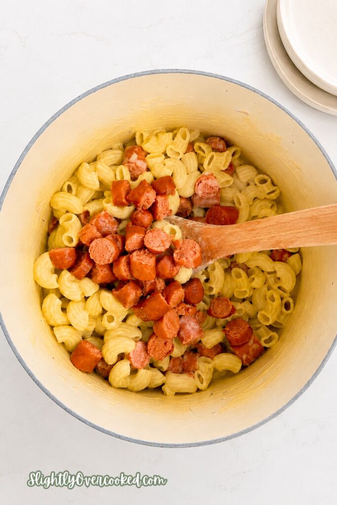 Hot dogs being cut into bite-sized pieces on a wooden cutting board with cooked macaroni nearby