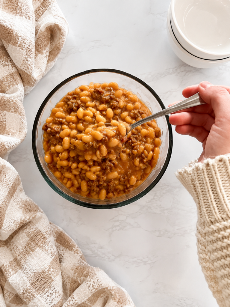 A spoonful of homemade baked beans with ground beef and bacon served in a bowl, featuring tender navy beans in a thick, flavorful sauce.