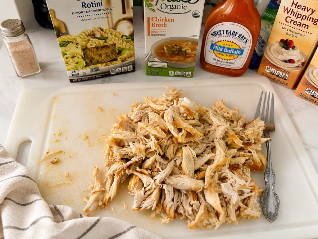 Shredded rotisserie chicken with rotini pasta, chicken broth, buffalo wing sauce, heavy cream, and seasonings prepared for an easy one-pot dinner recipe.