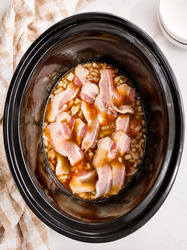 Slow cooker baked beans assembled with soaked navy beans, onion, ground beef, and bacon layered on top before slow cooking.