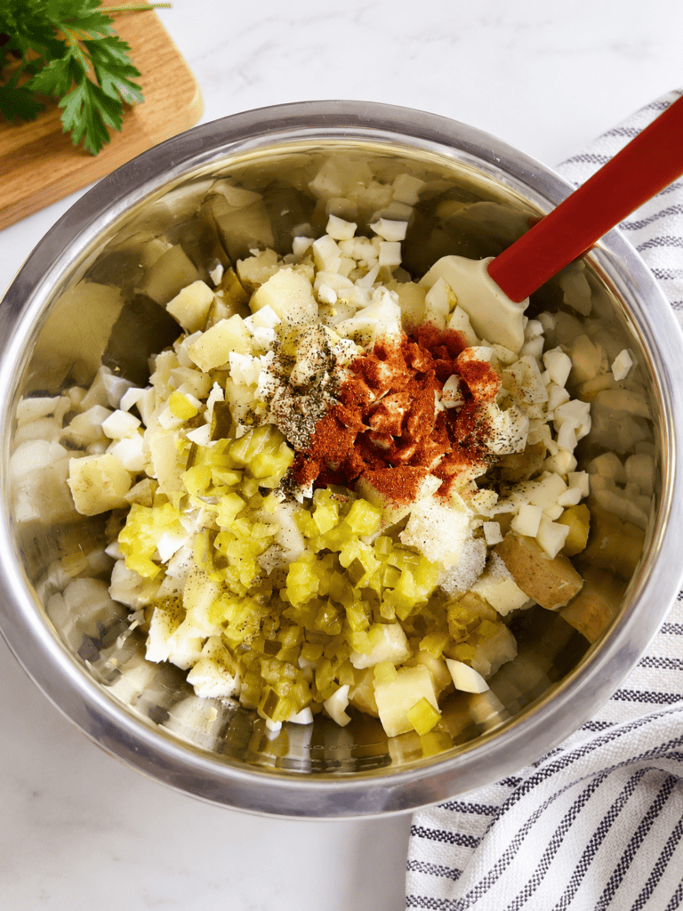 Ingredients for creamy white potato salad in a large mixing bowl including cooked Yukon Gold potatoes, chopped hard-boiled eggs, dill pickles, paprika, black pepper, salt, and mayonnaise before mixing. Easy classic potato salad recipe made with simple ingredients for a delicious homemade side dish.