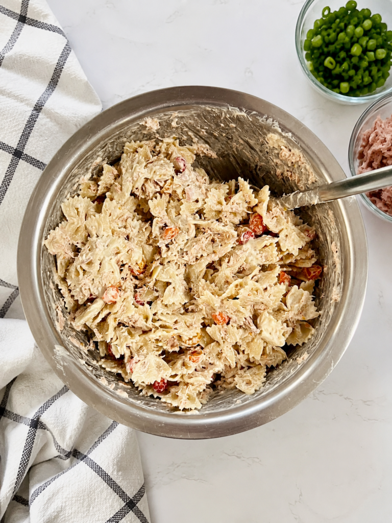 Creamy tuna pasta salad with bowtie pasta, canned tuna, cherry tomatoes, and mayonnaise mixed in a large stainless steel bowl on a white countertop with fresh green onions and a kitchen towel.