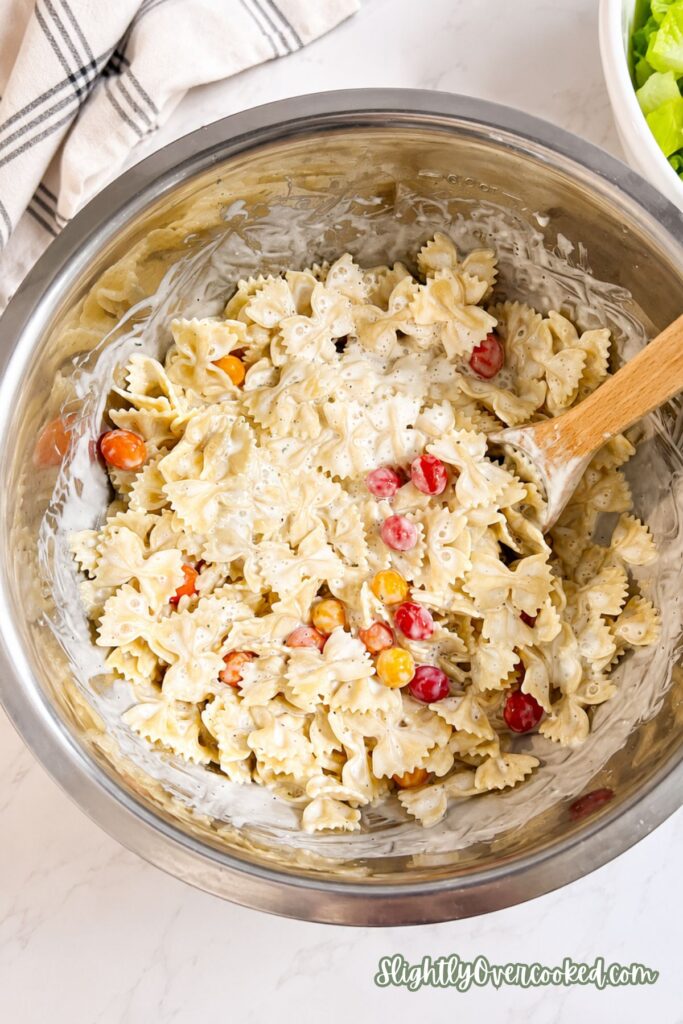 Cherry tomatoes being added to creamy bow tie pasta before mixing with bacon and lettuce.