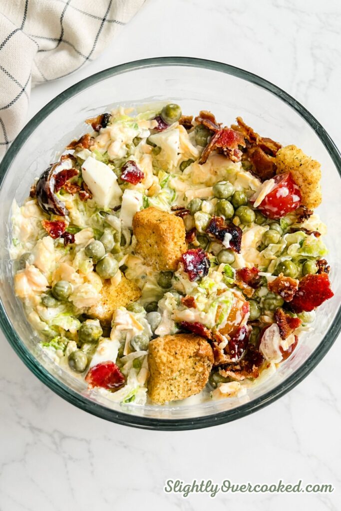 Close-up view of colorful layers of peas, cheddar cheese, bacon, tomatoes, lettuce, and creamy dressing in a glass bowl