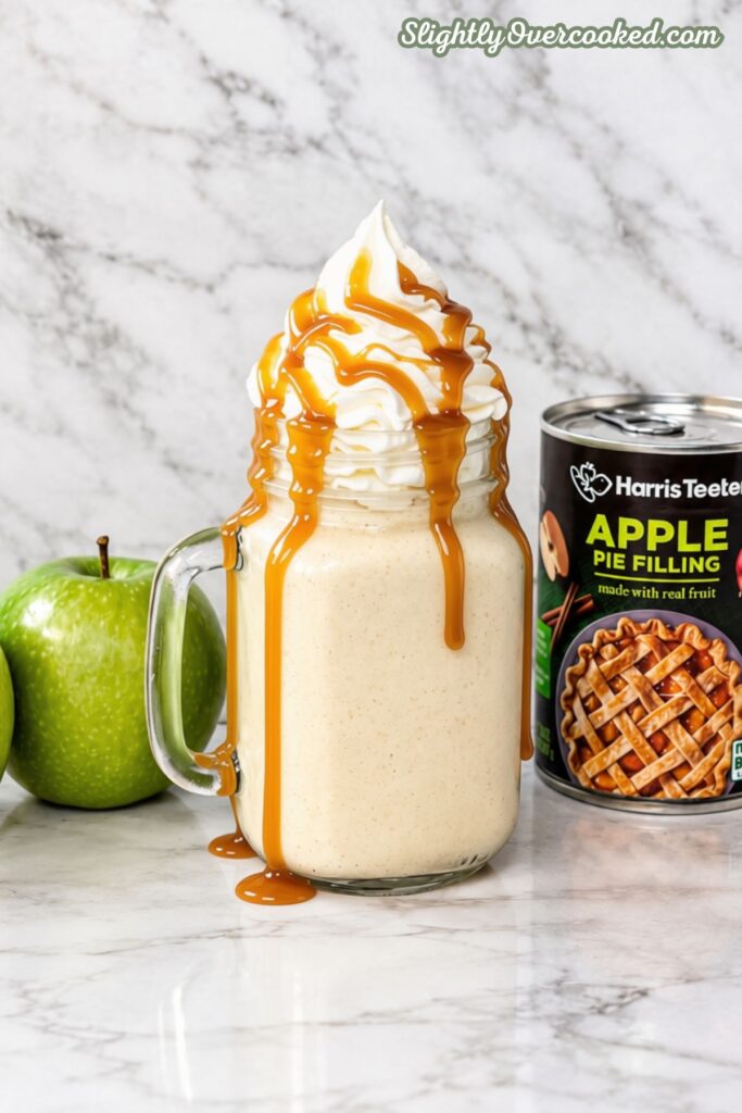 sitting on a marble counter top, this is a apple pie Milkshake in a mason jar glass with a handle. Topped with tall whip cream and drizzled with caramel sauce. Behind it is a can of apple pie filling and a green apple.