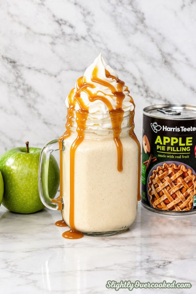 sitting on a marble counter top, this is a apple pie Milkshake in a mason jar glass with a handle. Topped with tall whip cream and drizzled with caramel sauce. Behind it is a can of apple pie filling and a green apple.