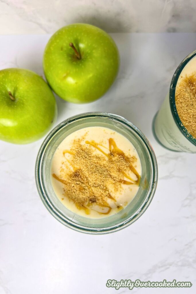 This is a apple pie milkshake in a plain glass with graham cracker crumber on top and caramel drizzle. its supper simple and doesn't have to be fancy. There is also two green apples in the background.